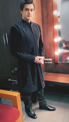 a man standing in front of a mirror wearing a black suit and tie with his hand on his hip