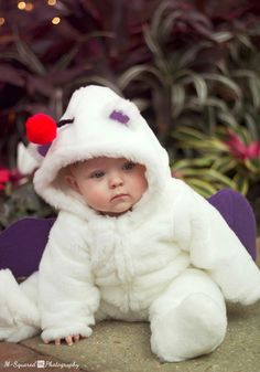 a baby dressed in a white costume sitting on the ground next to some plants and flowers