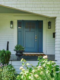 a blue front door on a white house with flowers in the foreground and bushes to the side