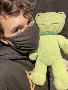 a man wearing a face mask and holding a green stuffed frog in front of him