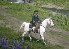 a woman riding on the back of a white horse next to a river and purple flowers