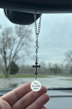 a person holding up a small cross on a car's front window with the words this too will pass written on it