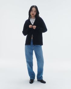 a woman standing in front of a white background wearing blue jeans and a black cardigan