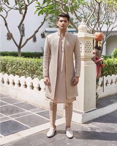 a man standing in front of a white fence wearing a beige outfit and shoes with his hands on his hips