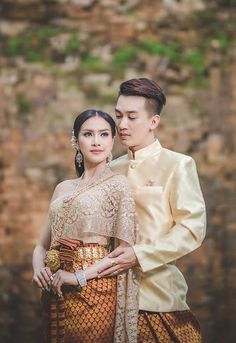 a man and woman standing next to each other in front of a stone wall wearing traditional thai garb