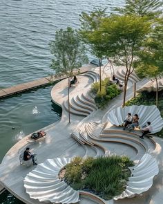 people sitting on benches near the water in an open area with trees and plants around them