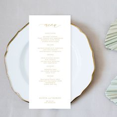 a white and gold place setting with menu cards on the plate, along with seashells