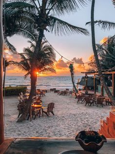 the sun is setting on the beach with palm trees and chairs in front of it