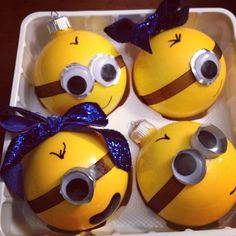 four yellow and brown ornaments in a plastic container with blue ribbon on them, decorated like minions from despicables