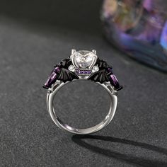 A cool black bat ring boasts a central, dazzling round gemstone, with bat images sculpted on both sides, infusing a touch of the macabre and the enchanting. Surrounding the main stone are sparkling purple and white stones, creating a striking and elegant contrast. Crafted from polished silver metal, the two tone fashion black ring features intricately carved black bat wing decorations, thereby enhancing its mystical charm. Inside, a spider's web-like hollowed-out lining lends an air of whimsy. It's a perfect fusion of sophistication, mystery, and allure, making it a standout accessory for any occasion.Carat Weight: 3.5 ctStone Size: 8 mmStone Type: Jeulia® StoneNumber of Stones: 1 Stone Color: Diamond WhiteStone Shape: RoundCarat Weight: 2.254 ctStone Size: 1.5,1,5*7 mmStone Type: Jeulia® Black Fantasy Rings As Gift, Black Fantasy Style Rings As Gift, Fantasy Style Black Ring For Gift, Elegant Black Skull Ring For Halloween, Bat Images, Bat Ring, White Stones, Bat Wing, Black Ring