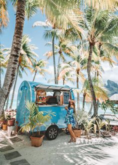 a woman standing in front of a blue food truck on the beach with palm trees