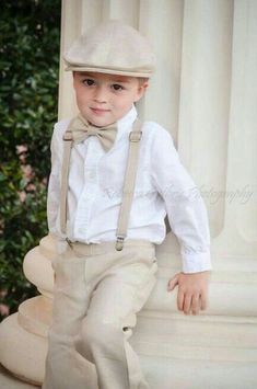 a young boy in a white shirt and bow tie standing next to a pillar wearing suspenders and a hat