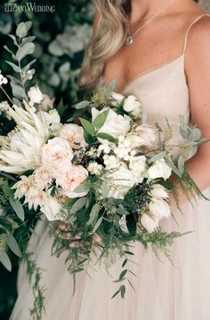 a woman holding a bouquet of flowers and greenery in front of her face, wearing a wedding dress