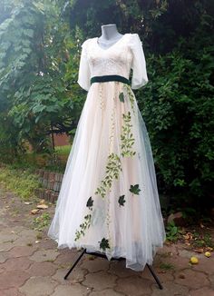 a white dress with green leaves on it sitting on top of a wooden stand in front of some bushes