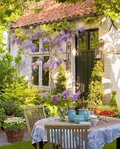 a table with two vases on it in front of a white building and purple flowers