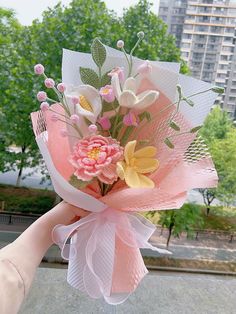 a hand holding a bouquet of flowers in front of a building with trees and buildings behind it