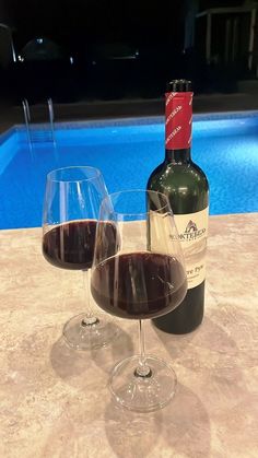 two wine glasses sitting next to each other on a table near a swimming pool at night