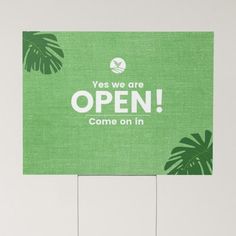 a green sign that says yes we are open come on in with palm leaves around it