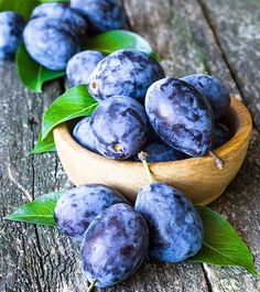 a wooden bowl filled with blue plums next to green leaves on top of a wooden table