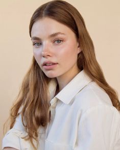 a woman with long brown hair wearing a white shirt and looking at the camera while standing in front of a beige wall