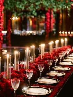 a long table with many plates and wine glasses on it, surrounded by red flowers