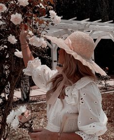 a woman in a white dress and straw hat picking flowers from a tree with her hand