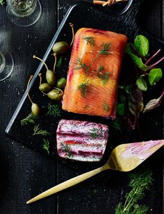 an assortment of raw fish and vegetables on a black platter with spoons next to it