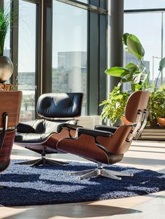 an eames lounge chair and ottoman in front of large windows with potted plants