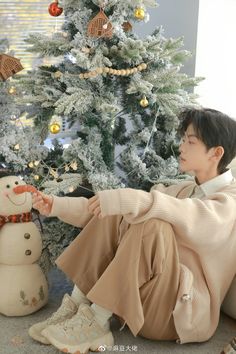 a young man sitting in front of a christmas tree with a snowman next to him