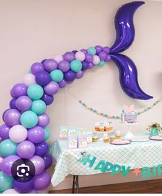 a birthday party with balloons, cake and mermaid tail decorations on a table in front of a balloon arch