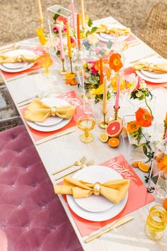 the table is set with yellow and pink plates, silverware, orange napkins, and gold place settings