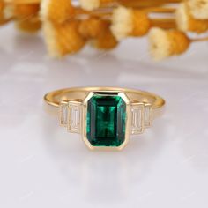 an emerald and baguette cut diamond ring on a white surface with dried flowers in the background