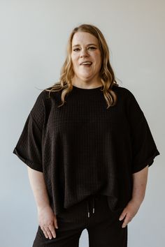 A person with long hair stands against a plain white background, wearing Mikarose's textured oversized tee with rolled cuffs and a scooped neckline, along with matching pants. Relaxed Fit Waffle Knit Crew Neck Top, Casual Black Waffle Knit Top, Textured Crew Neck Tops For Fall, Textured Crew Neck Top For Layering, Knit Textured Tops For Layering, Black Ribbed Neckline Top For Loungewear, Textured Casual Tops For Loungewear, Black Knit Tops For Loungewear, Relaxed Fit Short Sleeve Waffle Knit Tops