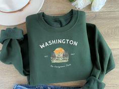 This high quality, sustainably made Washington State crewneck is the coziest gift to remember all your adventurous memories of the Evergreen State. Featuring the classic phrase "WASHINGTON est 1967 The Evergreen State," this crewneck is perfect for anyone who loves Washington's natural beauty and laid-back vibes. Crafted from 80% ring spun cotton and 20% polyester, the Gildan® SF000 sweatshirt offers a super soft feel with its brushed interior and 100% ring spun cotton face. The medium-heavy fabric (8.4 oz/yd²) makes it perfect for chilly days or cozy nights. With its dropped shoulder design for a relaxed fit, this crewneck will quickly become your go-to. We've made sure that style and comfort align with eco-conscious production. The sweatshirt is made with OEKO-TEX certified low-impact dy Green Fleece Sweatshirt For Outdoor, Green Fall Sweatshirt For Outdoor Activities, Green Sweatshirt For Fall Outdoor Activities, Green Sweatshirt For Outdoor Fall Activities, Green Winter Sweatshirt For Outdoor Activities, Green Crew Neck Sweatshirt For Outdoor, Green Cotton Outdoor Sweatshirt, Green Cotton Sweatshirt For Outdoor, Green Fleece Tops For Outdoor Activities