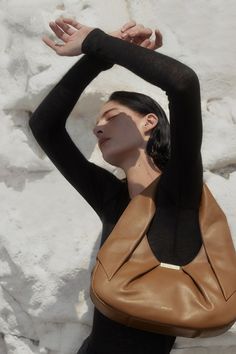 a woman with her hands behind her head, leaning against a wall holding a brown purse