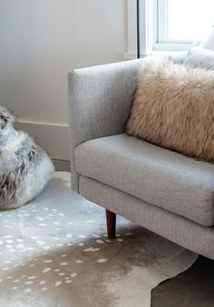 a gray couch sitting on top of a white rug next to a window with a fur pillow