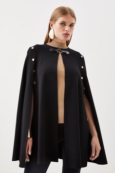 a woman is wearing a black cape with buttons on the front and side, she has her hands in her pockets