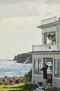 two people standing on the porch of a white house by the ocean with trees and water in the background