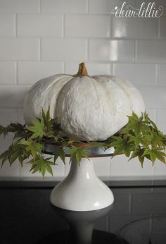 two white pumpkins sitting on top of a cake stand with green leaves around them