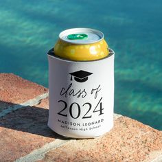 a can of beer sitting on top of a rock next to the water with a graduation cap on it