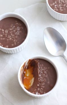 three white bowls filled with chocolate pudding on top of a table next to two spoons