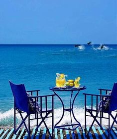 two chairs and a table with drinks on it near the water's edge in front of an ocean