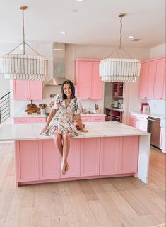 a woman sitting on top of a pink island in a kitchen with chandeliers