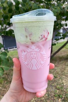 someone holding up a pink starbucks cup with strawberries and whipped cream on it in front of a tree