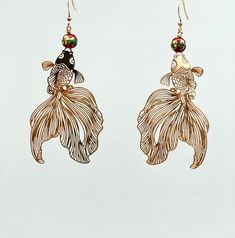 two gold fish earrings hanging from hooks on a white background, one with green and red beads