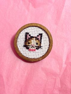 a small cross stitch cat on a pink background with a wooden frame for it's eyes