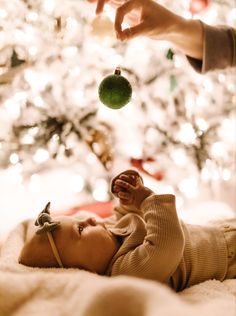 a baby laying in front of a christmas tree with a green ornament hanging from it's mouth