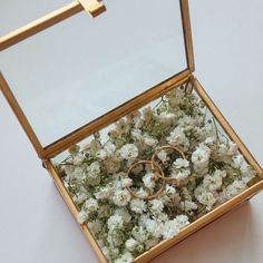 two gold wedding rings in a box filled with white flowers and greenery on a table