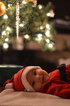 a baby wearing a santa hat laying on top of a blanket next to a christmas tree