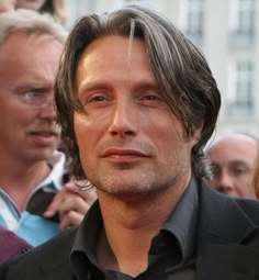 a man with long hair standing in front of a group of people and looking at the camera